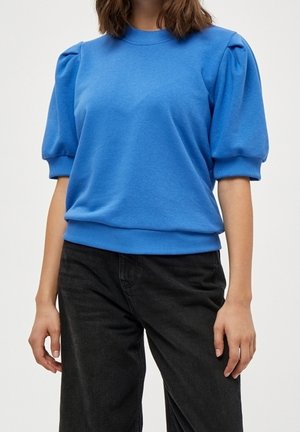 Frau trägt einen blauen Pullover mit Puffärmeln, gerippten Bündchen und Saum, kombiniert mit hoch taillierten schwarzen Hosen.