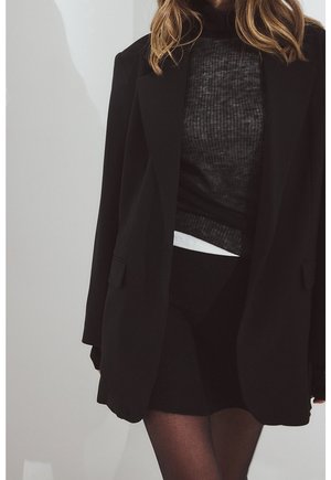 Kvinde iført en sort blazer over en grå ribbet sweater og sort nederdel, stående mod en lys grå baggrund.