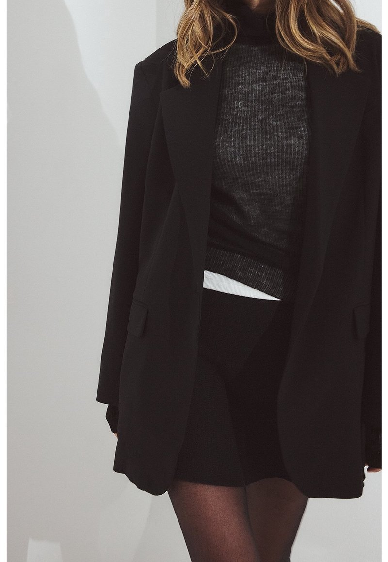Femme portant un blazer noir par-dessus un pull côtelé gris et une jupe noire, debout devant un fond gris clair.