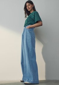 Camiseta verde a rayas combinada con unos vaqueros de denim azul claro de tiro alto y pernera ancha, que cuentan con bolsillos frontales y un dobladillo sin terminar.