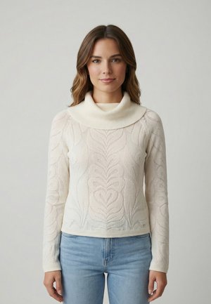 Mujer joven con cabello castaño que lleva un suéter de cuello alto color crema con estampado y jeans azul claro, de pie contra un fondo liso y claro.