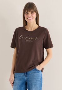 Femme souriante portant un t-shirt marron avec le texte "Curieuse par nature, va où tu te sens le plus vivante", associée à un jean bleu, main dans la poche.