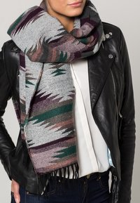 Écharpe grise tissée avec des motifs géométriques multicolores en vert, marron et violet, avec des bords frangés, drapée autour d'une veste en cuir noire.