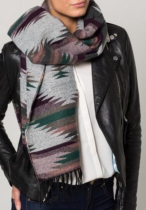 Écharpe grise tissée avec des motifs géométriques multicolores en vert, marron et violet, avec des bords frangés, drapée autour d'une veste en cuir noire.