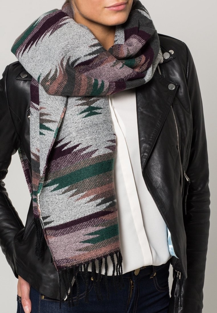 Écharpe grise tissée avec des motifs géométriques multicolores en vert, marron et violet, avec des bords frangés, drapée autour d'une veste en cuir noire.