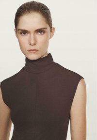 Haut marron sans manches à col montant avec une silhouette ajustée et une texture lisse, présentant un détail subtil de couture. Fond blanc uni.