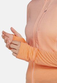 Veste légère à manches longues zippée de couleur orange clair. Elle est fabriquée dans un tissu doux et texturé avec des passe-pouces et une fermeture éclair à l'avant.