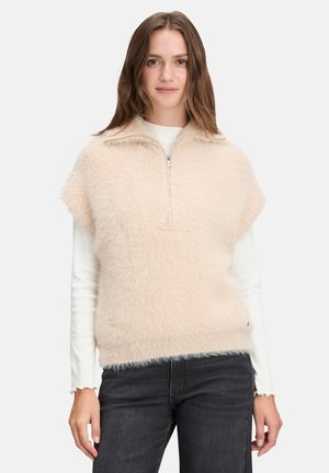 Gilet en fausse fourrure rose clair avec un zip sur le devant et un col fendu, porté par-dessus une chemise blanche à manches longues, associé à un jean sombre.