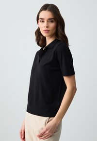 Polo shirt nero a maniche corte realizzato in tessuto morbido, con colletto e una sottile texture a coste all'orlo. Indossato con pantaloni chiari.