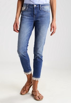 Femme portant un jean slim bleu clair avec une bordure en dentelle blanche aux chevilles et un petit détail en dentelle sur la poche avant, associé à des sandales marron.