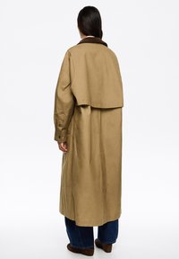 Chaki trenčkotas su velveto apykakle, oversized siluetu, ilgomis rankovėmis ir mygtukais uždaromais манжетами. Yra nugaros ventiliacija judėjimui.
