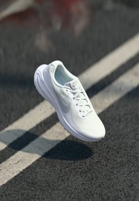 White lightweight running shoe with mesh upper, padded collar, and rubber sole. Features grey Nike logo and laces, suspended above pavement.