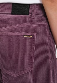 Pantalon en velours côtelé violet avec des rainures verticales, équipé d'une étiquette noire sur la ceinture et d'une petite  étiquette dorée "VOLCOM" sur la poche arrière.