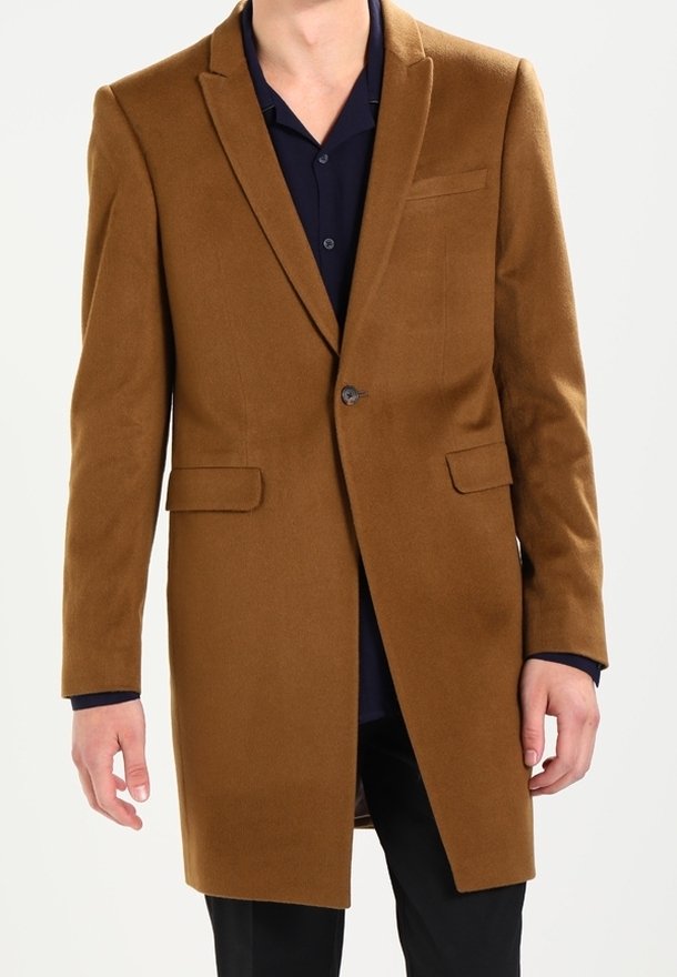Homme portant un manteau marron à un bouton sur une chemise bleu marine foncé avec un pantalon noir, debout devant un fond uni clair.