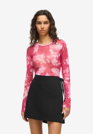 Kvinde med krøllet hår iført en langærmet lyserød tie-dye top og et sort omslagsskjørt, der står foran en hvid baggrund.