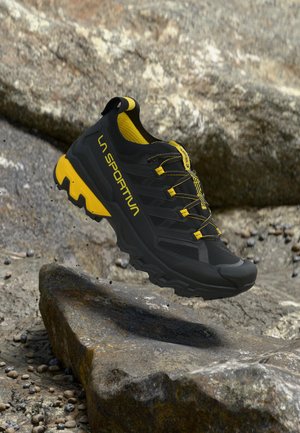 Zwart-gele La Sportiva trailrunningschoen in de lucht boven rotsachtig terrein met aarde en kleine stenen verspreid eromheen.