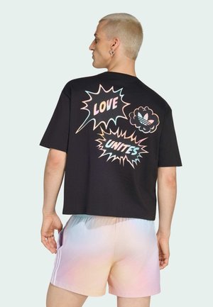 Mężczyzna z krótkimi blond włosami, ubrany w czarną koszulkę z pastelowym napisem "LOVE UNITES" i logo, w połączeniu z różowo-niebieskimi, gradientowymi szortami.