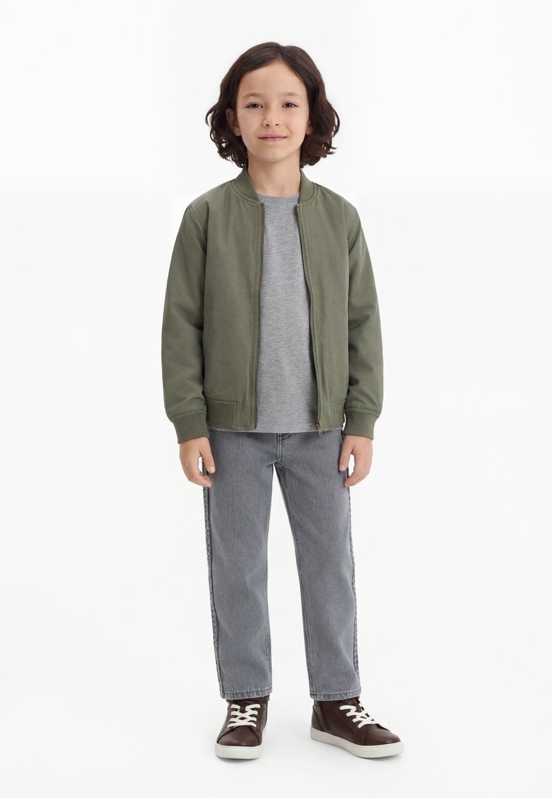Gyermek áll, zöld bomberkabátot, szürke pólót, szürke farmert és barna sportcipőt visel fehér fűzővel, egyszínű fehér háttér előtt.