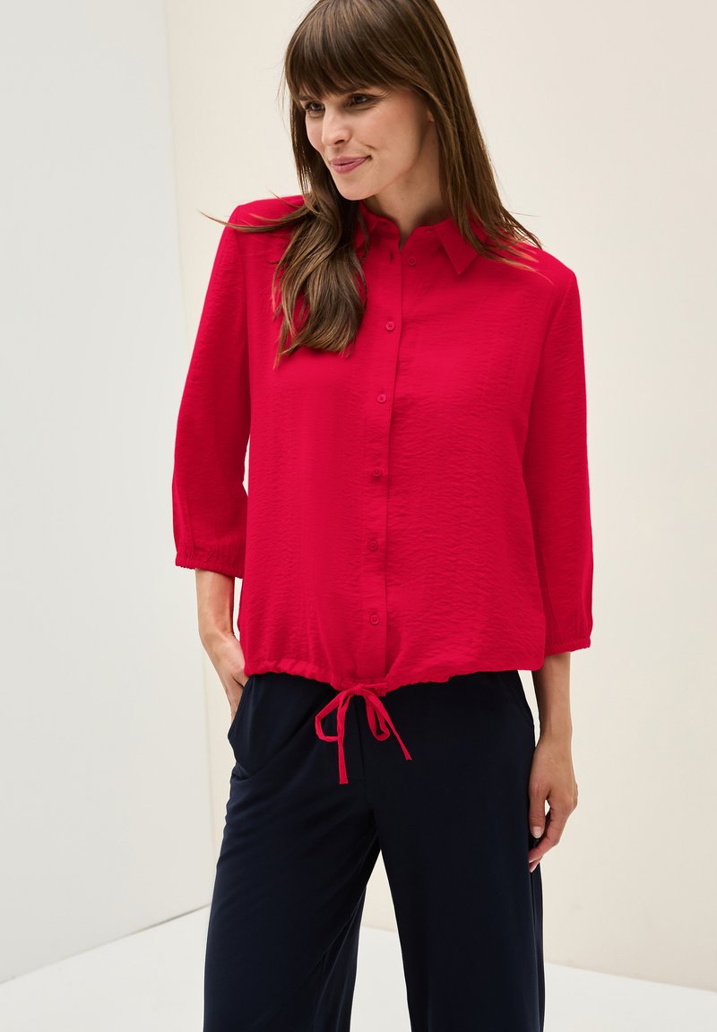 Chemise rouge à manches trois-quarts, en tissu léger, surface texturée et ourlet à cordon, associée à un pantalon large et sombre.