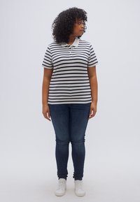 Ριγέ πόλο μπλουζάκι σε γκρι και ναυτικό μπλε με γιακά, συνδυασμένο με σκούρο μπλε στενά τζιν και λευκά αθλητικά παπούτσια, ποζάροντας μπροστά σε απλό φόντο.