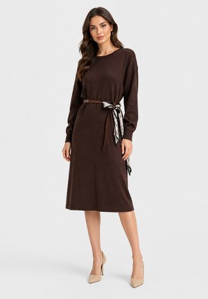 Donna che indossa un abito midi marrone scuro a maniche lunghe con una cintura sottile e una sciarpa a fantasia annodata in vita, abbinato a décolleté beige.