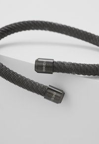 Accessorio in filo a spirale nero con estremità in metallo cromato. Le estremità presentano il marchio "BREIL" inciso. La texture è liscia e il design è flessibile.
