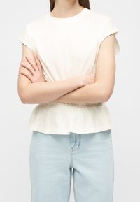 Femme les bras croisés portant un chemisier blanc à manches courtes avec un bas peplum et un jean taille haute bleu clair.
