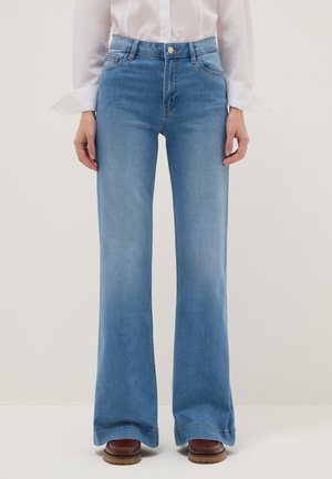 Vaqueros acampanados de cintura alta en azul claro, combinados con una camisa blanca de manga larga y zapatos con suela marrón, de pie contra un fondo liso.