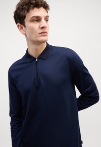 Polo de manga longa azul-marinho com colarinho com zíper, apresentando um padrão texturizado e um design ajustado. Etiqueta de marca discreta na lateral.