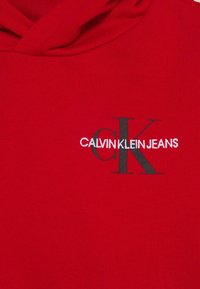 Felpa rossa con cappuccio realizzata in tessuto morbido; presenta un logo "CK" nero e la scritta "CALVIN KLEIN JEANS" in bianco, centrata nella zona del petto.