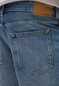 Jeanșii din denim albastru au o buzăcă vizibilă pe spate cu cusături portocalii și o etichetă din piele bej marcată cu "PULL&BEAR" la talie.