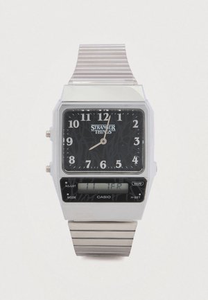 Orologio digitale Casio con cassa rettangolare argentata, quadrante nero che mostra la scritta "STRANGER THINGS", cinturino metallico argentato e display doppio orario analogico-digitale.