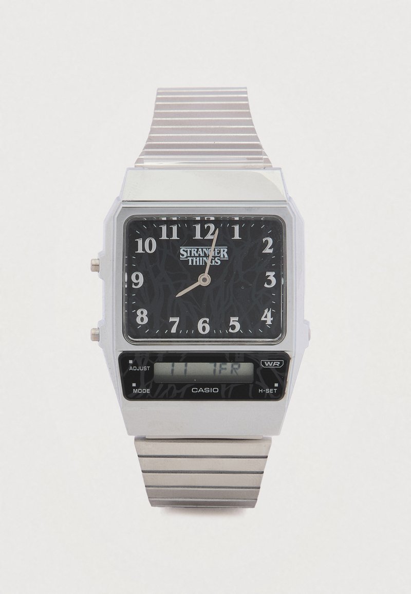 Montre numérique Casio avec un boîtier rectangulaire en argent, cadran noir affichant "STRANGER THINGS", bracelet en métal argenté, et affichage dual temps analogique-numérique.
