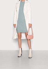 Cappotto lungo bianco, vestito lavorato a maglia azzurro chiaro, borsa rosa, stivaletti beige alla caviglia con accenti neri e punte argentate. Texture lisce e design moderno.