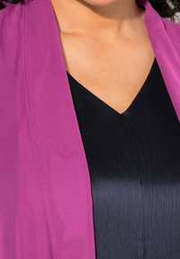 Kvinna i en svart V-ringad topp under en magenta blazer, som visar halslinjen och övre bröstområdet.