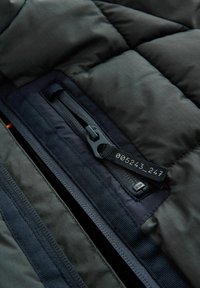 Gros plan d’une fermeture éclair d’une veste matelassée noire avec une languette en tissu indiquant des chiffres et un tissu matelassé texturé autour.