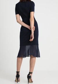 Vestido navy con mangas cortas, cuerpo ajustado y un dobladillo acampanado y transparente. Usado con zapatos de tacón alto negros, mostrando la vista trasera.