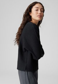 Schwarzer, strukturiert gestrickter Pullover mit lockerem Schnitt, weitem Ärmel, rundem Ausschnitt und seitlichen Schlitzen. Kombiniert mit grauer Hose.