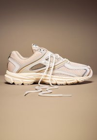 Baskets de sport beige et crème avec des détails en mesh et des lacets blancs défaits, positionnées sur une surface et un fond beige assortis.