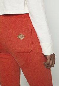 Červené tepláky vyrobené z měkké tkaniny mají zadní kapsu s texturovanou, kosočtvercovou nášivkou loga. Pas je elastický.