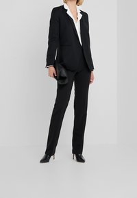 Zwarte op maat gemaakte pak met een witte overhemelse, met een getailleerd blazer, strakke broek en elegante zwarte hakken. Houdt een zwarte clutch vast.