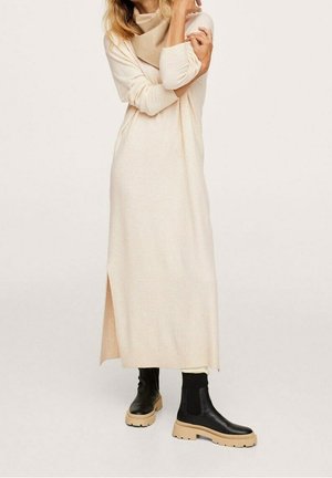 Vrouw die een lange beige sweaterjurk met zijsplit draagt en zwarte laarzen met dikke beige zolen, armen gekruist op de borst.