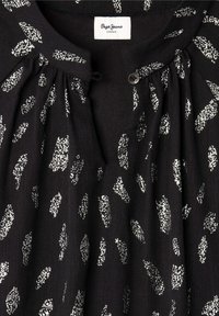Blusa negra con un patrón salpicado de plata, que presenta un escote redondo y botones decorativos. La tela tiene un aspecto ligero y texturizado.