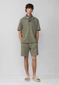 Olivgrünes kurzärmeliges Hemd mit Knopfleiste und passende Shorts aus strukturiertem Material, kombiniert mit beigen Slip-On-Sandalen und einem schwarzen gemusterten Schal.