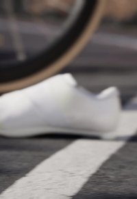 Scarpa atletica bianca con texture liscia, punta arrotondata e design minimalista, poggiata parzialmente su una linea bianca contro una superficie scura.