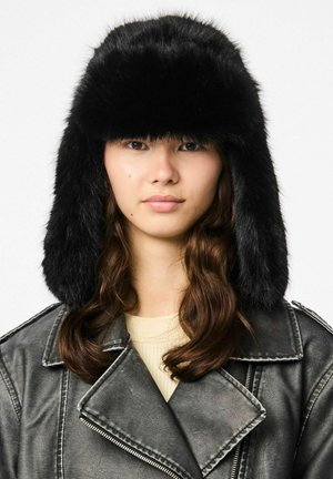 Sort faux fur trapperhat med øreklapper, der har en blød, fluffy tekstur og en rund top, båret af en model med langt, bølget hår.