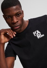 Zwarte katoenen t-shirt met ronde hals. Heeft een wit "KARL" logo op de borst. Model poseert met hand op kin, neutrale achtergrond.