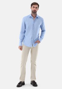 Camicia azzurro chiaro a maniche lunghe in tessuto morbido, con colletto a punta e bottoni frontali, abbinata a pantaloni beige e scarpe scure.