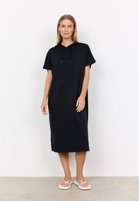 Svart oversized huvtröja med korta ärmar, med en framlåda, tillverkad av mjukt tyg med en slät yta. Bärs med lätta sandaler.