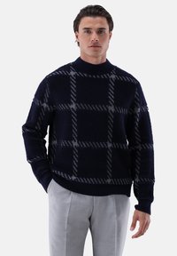 Marineblauer Pullover mit strukturiertem Strick, der ein graues Karomuster zeigt. Hoher Kragen und gerippte Bündchen, kombiniert mit hellgrauen Hosen.
